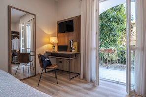 Habitación de hotel con un escritorio de madera, una laptop, una lámpara, una silla con almohada, un espejo y vistas a un balcón con vegetación.
