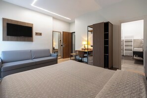 Habitación de hotel moderna con una cama grande, un sofá, un escritorio con lámpara, un armario y un baño visible a través de una puerta abierta.