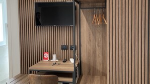 Een compacte, moderne kamer met een houten bureau, een stoel, een aan een gesloten wand bevestigde televisie en een kapstok.