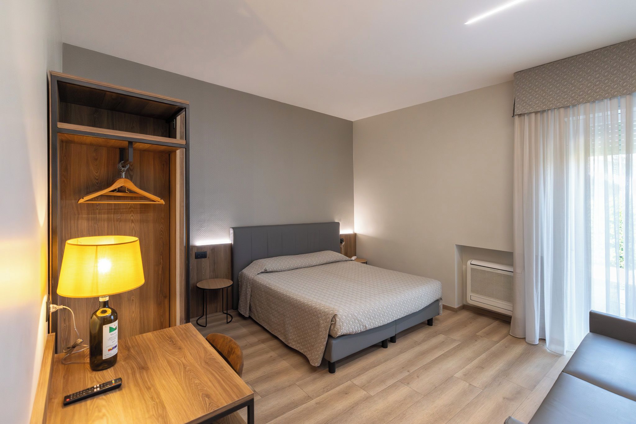 A hotel room with a bed, bedside table, lamp, and a wardrobe with a hanging garment.