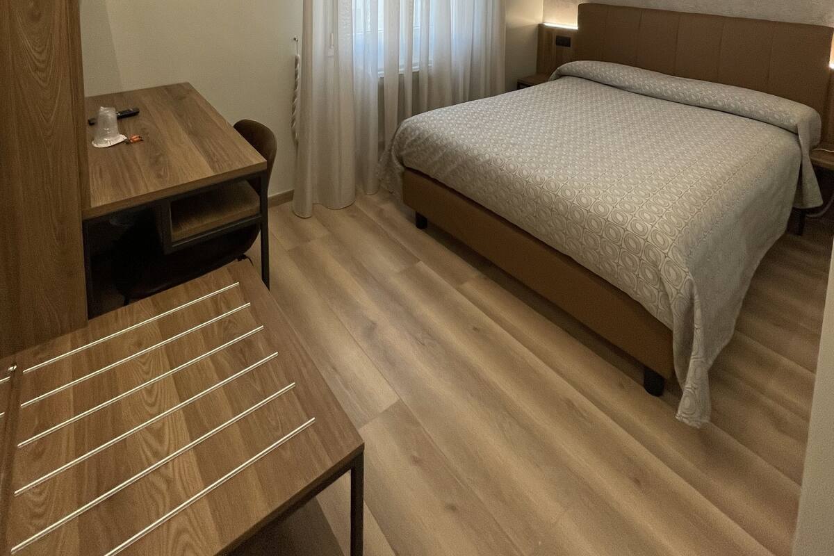 A hotel room with a wooden desk, a bed with a patterned bedspread, a nightstand, and a large window with sheer curtains.