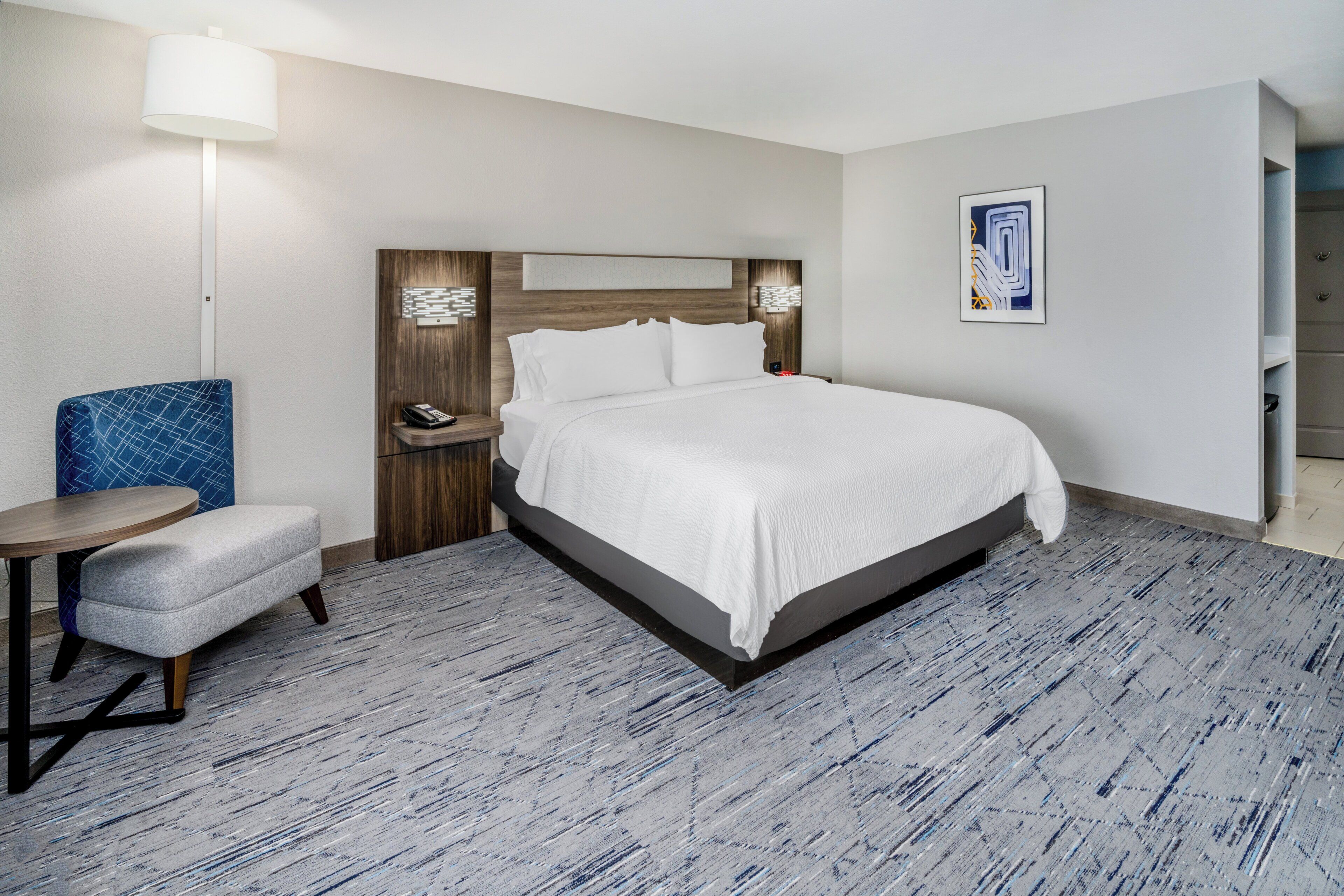 A hotel room with a large bed, a small round table, a blue chair, a bedside table with a lamp, and a framed picture on the wall.