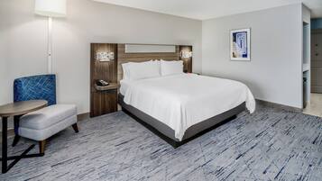 A hotel room with a large bed, a small round table, a blue chair, a bedside table with a lamp, and a framed picture on the wall.