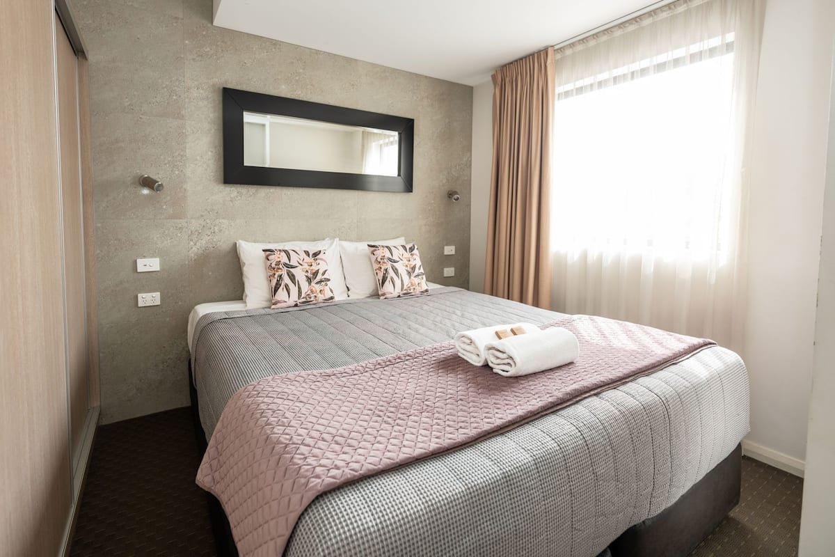 A hotel room with a bed, a large mirror, a window with curtains, and a wall with electrical outlets.