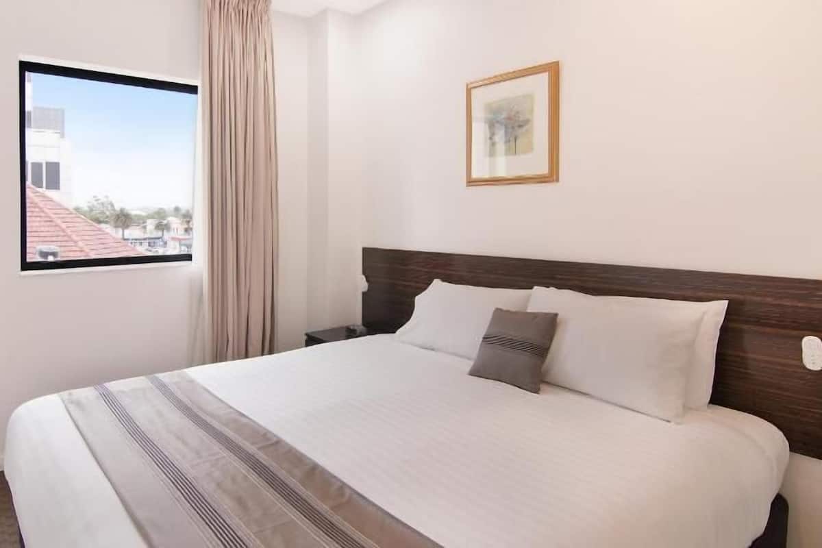 A hotel room with a large bed, a window with a view of buildings, a framed picture on the wall, and a nightstand with a lamp.