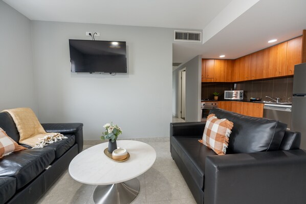 A modern living room with two black leather sofas, a white coffee table, and a flat-screen TV mounted on the wall.