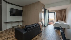 A modern hotel room with a black leather sofa, a flat-screen TV mounted on the wall, a wooden floor, a bed with a white mattress, and a sliding glass door leading to a balcony. - Boulevard on Beaumont (Hamilton)