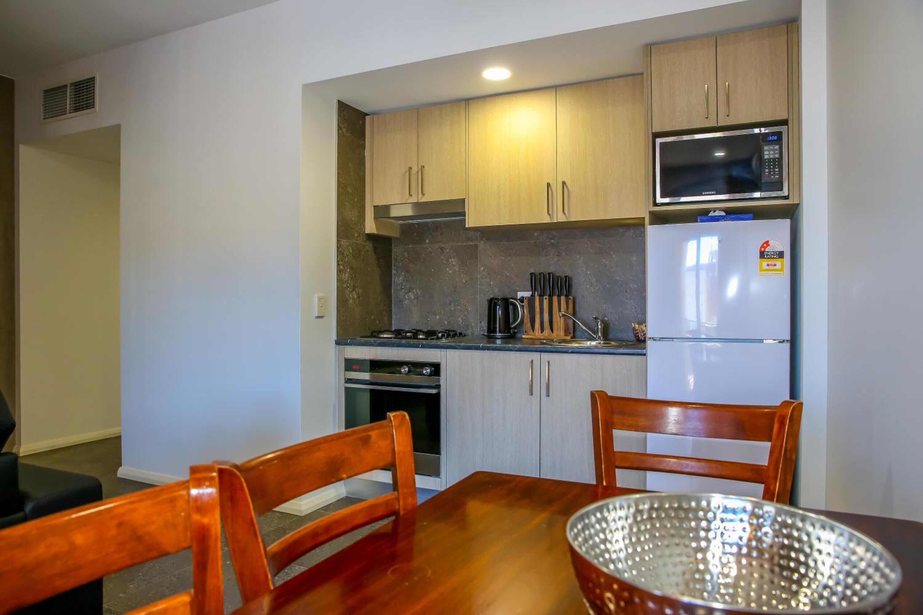 Cocina compacta con armarios de madera, microondas, refrigerador y una mesa de comedor con sillas.