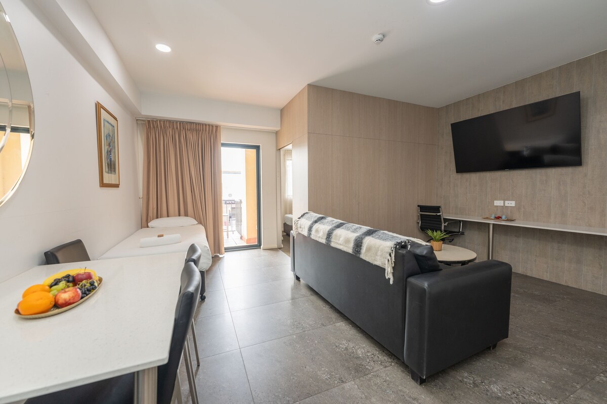A modern living room with a sofa, a dining table with fruit, a TV, and a view to the outside.
