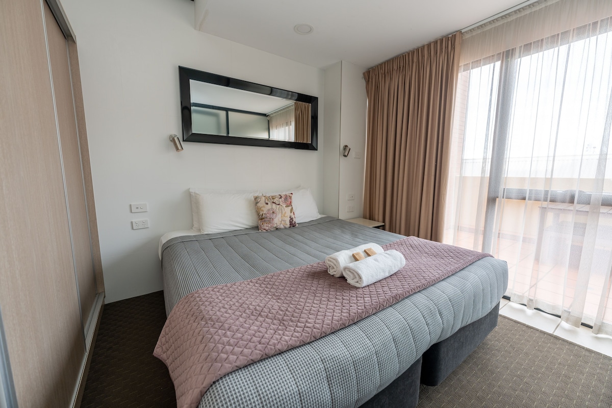 A hotel room with a large bed, a mirror, a window with curtains, and a view of the outdoors.