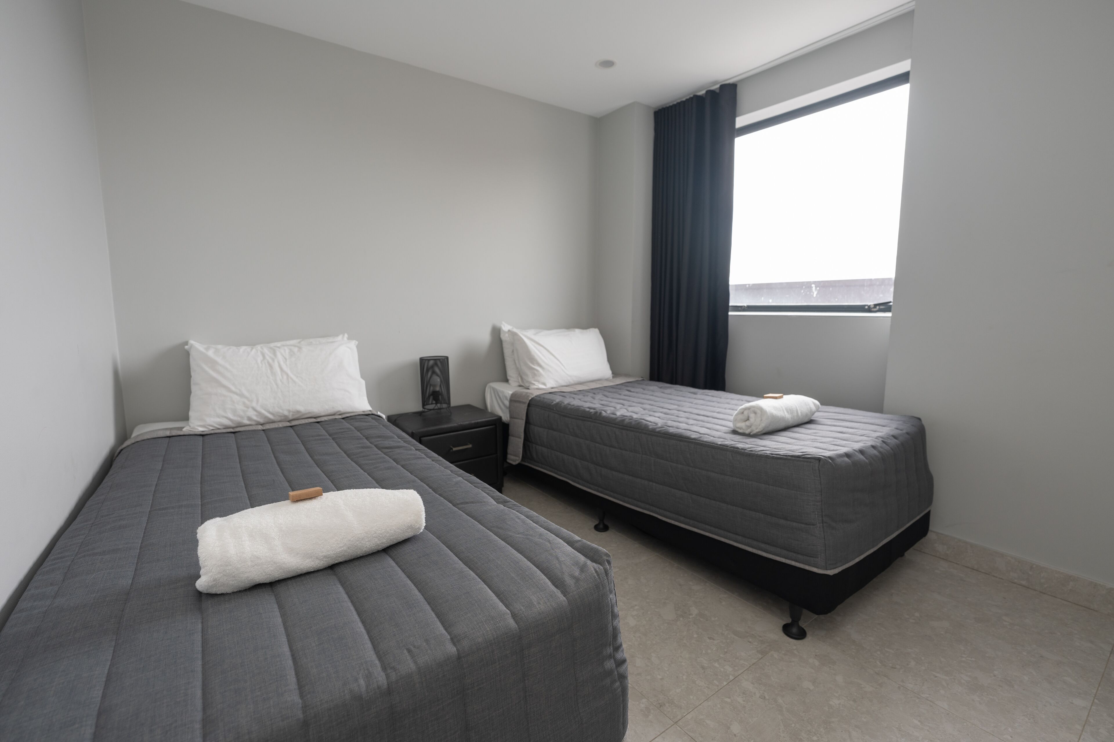 A modern hotel room with two beds, a nightstand, a window with curtains, and a view of the outside.