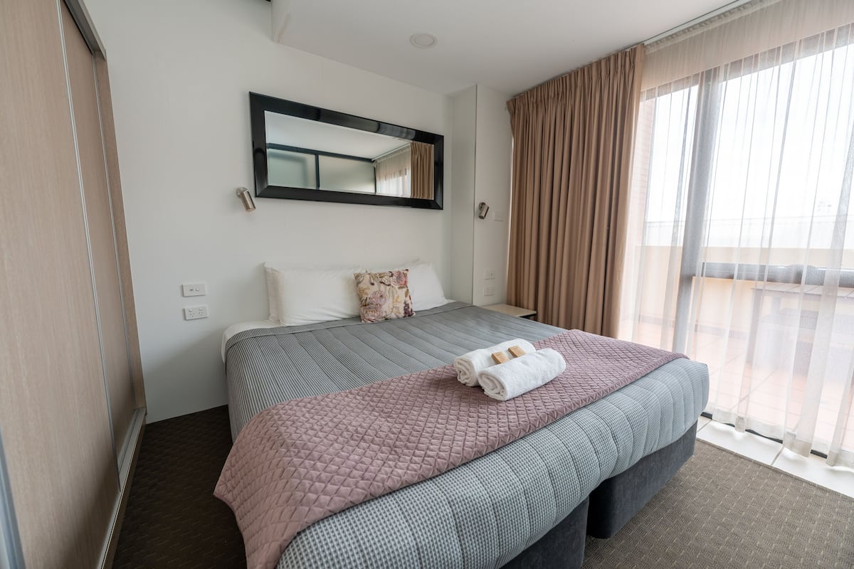 A hotel room with a large bed, a bedside table, a mirror, and a window with curtains.