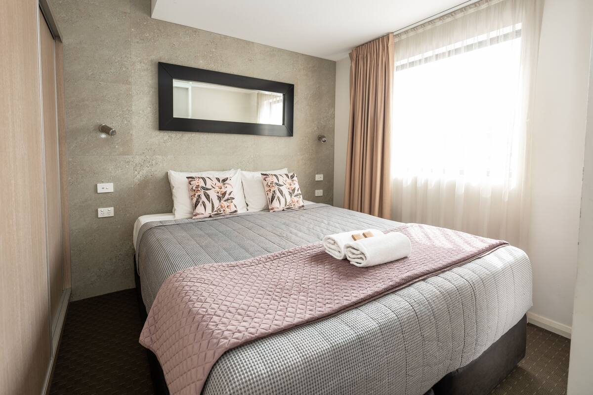 A hotel room with a large bed, a mirror above the bed, a window with curtains, and a wall-mounted light fixture.