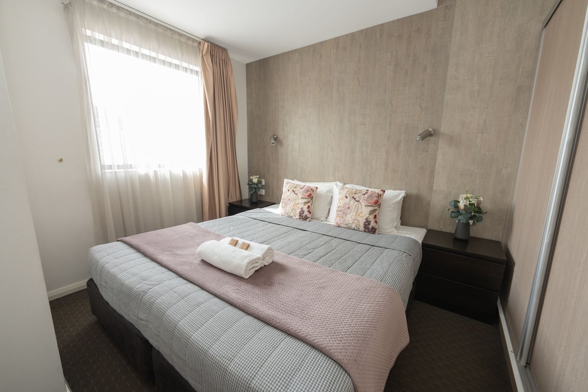 A neatly arranged hotel room with a large bed, a bedside table with a vase of flowers, and a window with curtains.