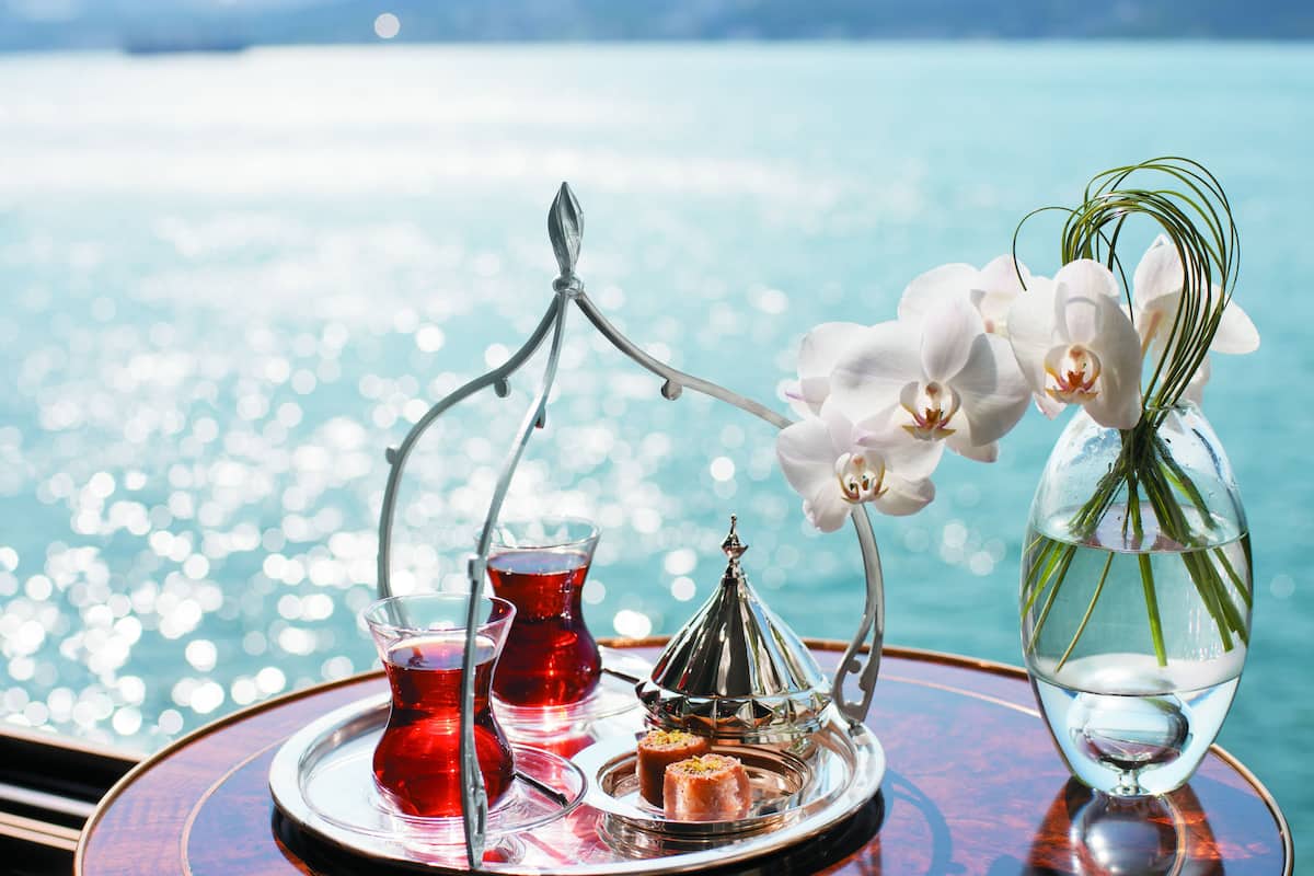 A tray with a glass of red tea, a silver teapot, and a small dish with food, set against a scenic view of water.