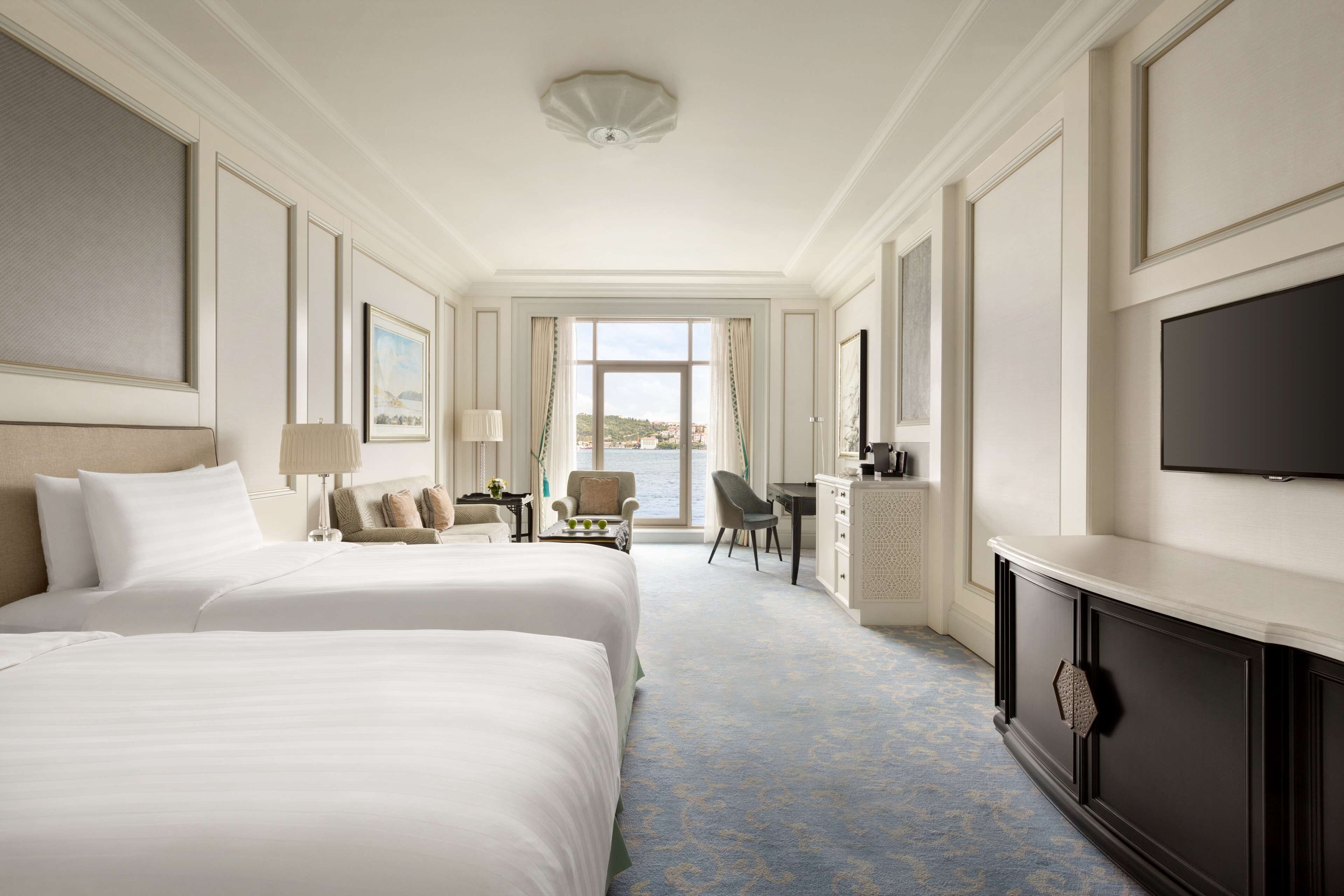 A hotel room with two large beds, a desk, a chair, a television, and a view of the city.