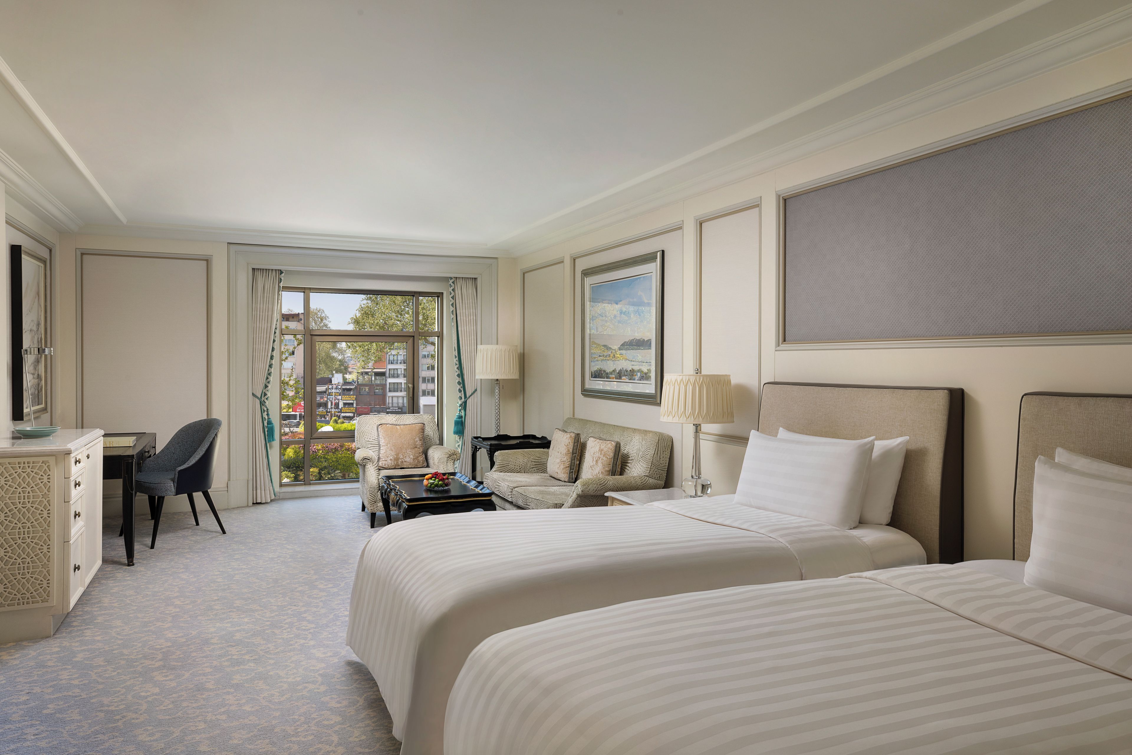 A hotel room with two beds, a desk, a chair, a small table with a bowl of fruit, and a view of the city through the window.