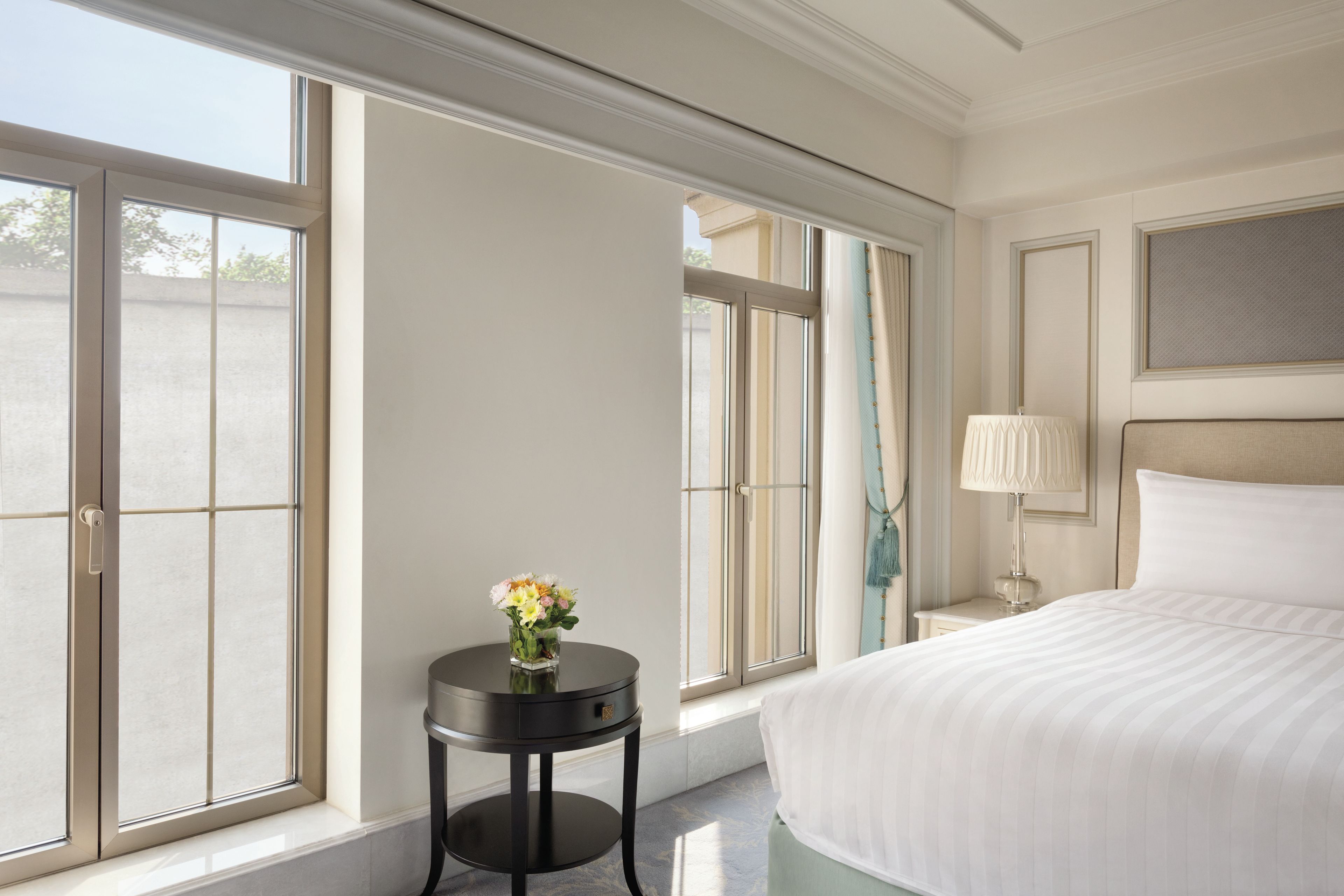 Een hotelkamer met een groot bed, een nachtkastje met een lamp, een klein rond tafeltje met een bloemenvaas, en uitzicht door het raam.
