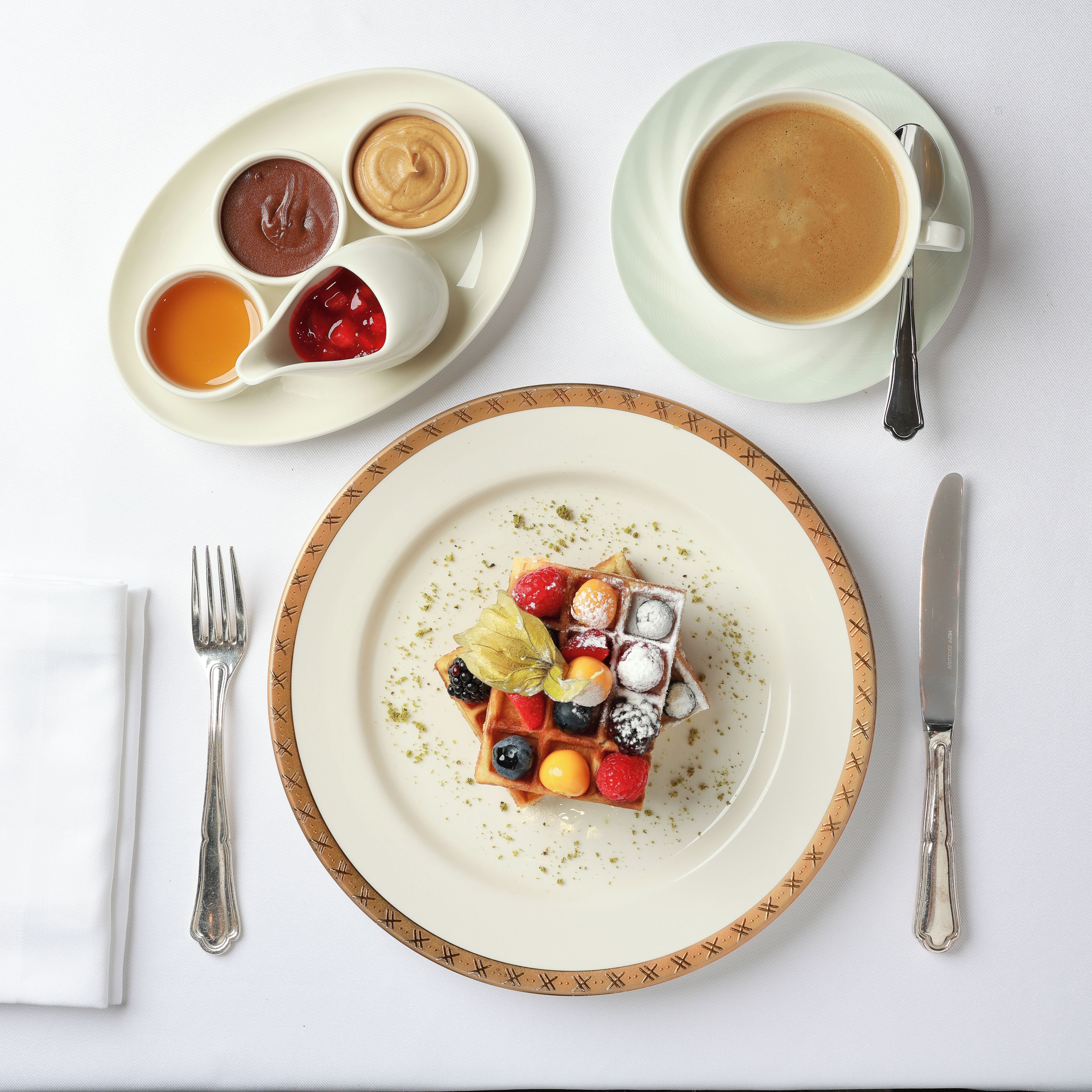A breakfast setting with a plate of waffles topped with berries and powdered sugar, a cup of coffee, and two small bowls of jam and honey.