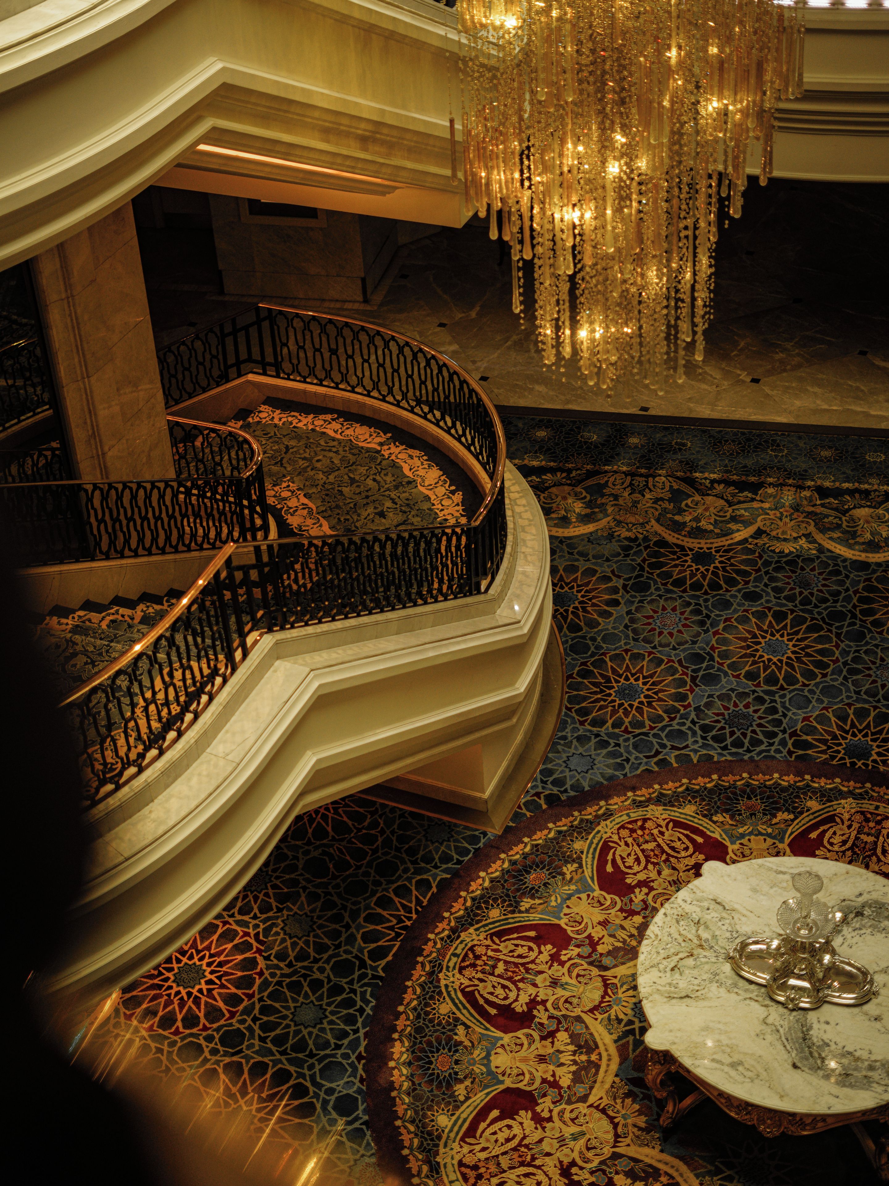 A grand interior with a marble staircase, ornate carpet, and a chandelier.