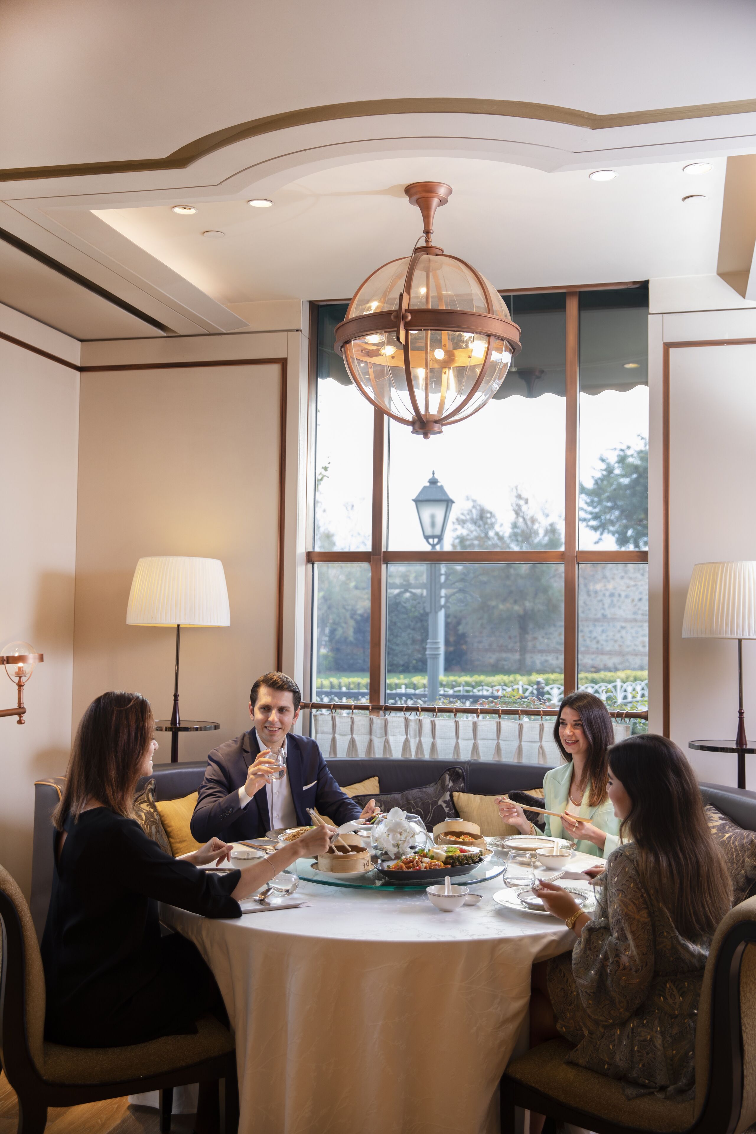 A dining area with a round table, four people seated, and a large window with a view of greenery.