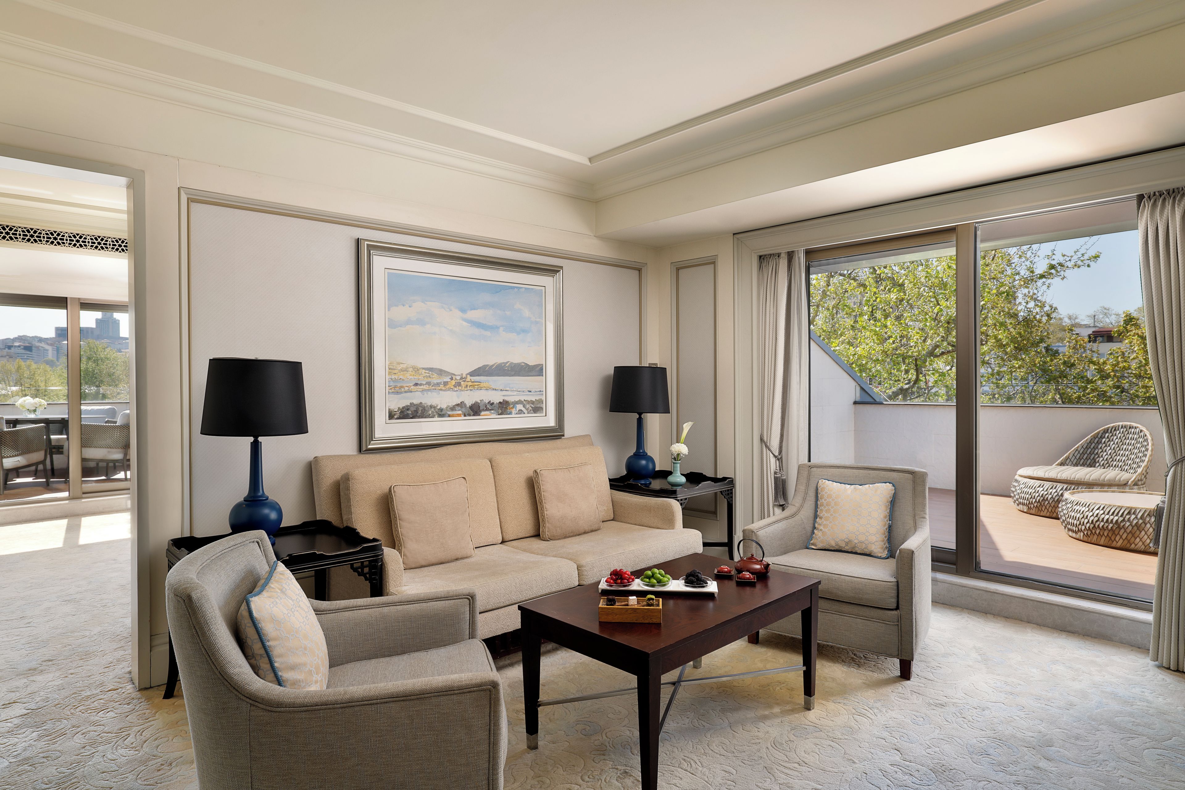 A well-lit living room with a beige sofa, two armchairs, a coffee table with decorative items, and a large window with a view of greenery.