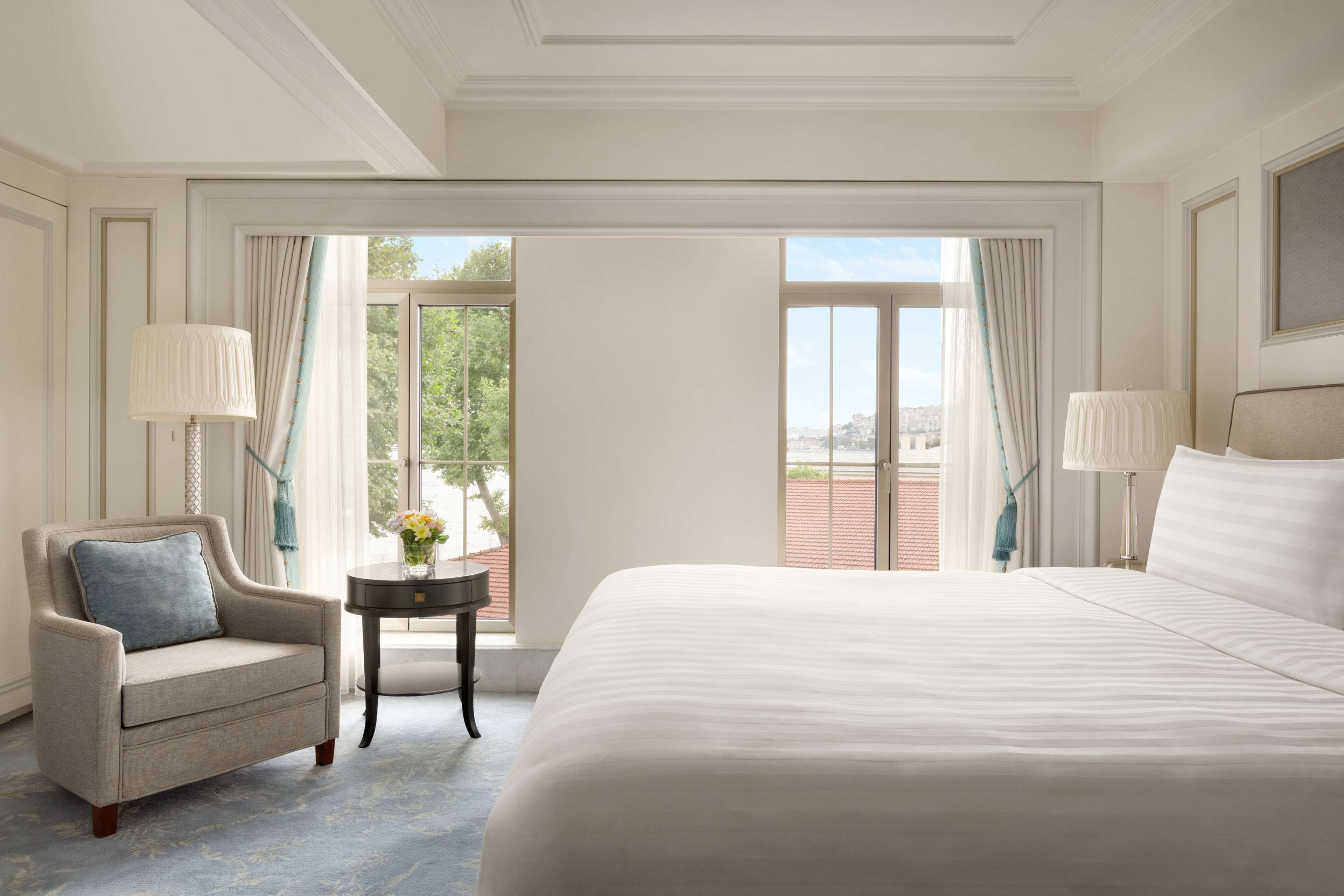 A spacious bedroom with a large bed, two bedside lamps, a small table with a vase, and a view of the outdoors through a large window.