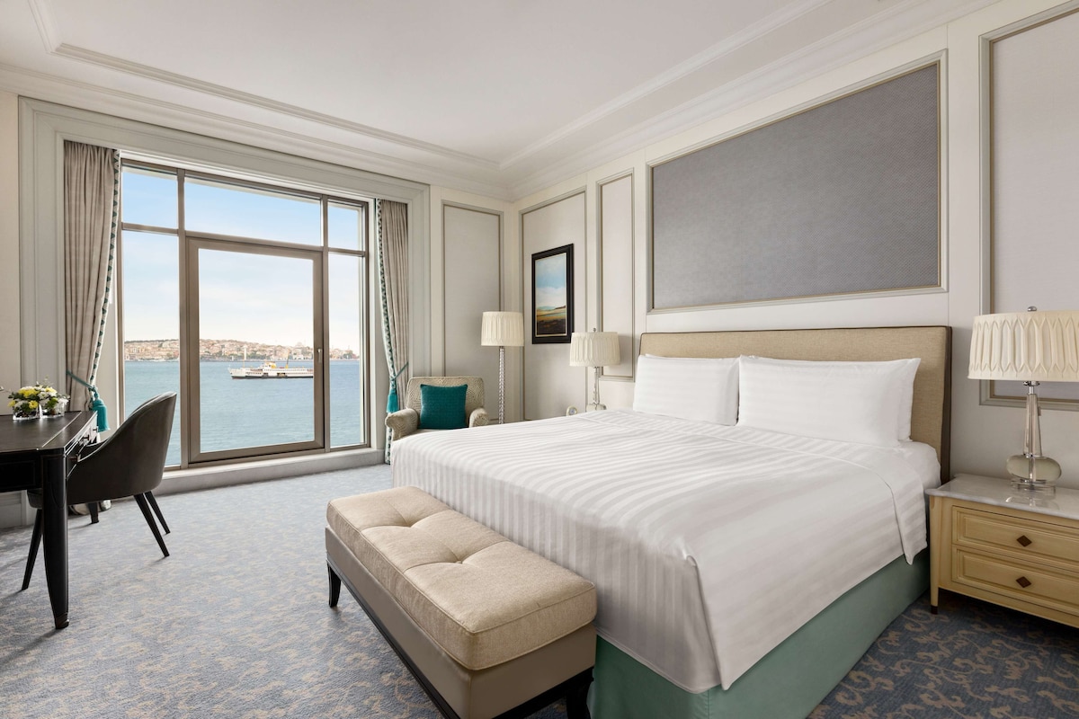 A hotel room with a large bed, a desk, a chair, a bench, and a view of the sea.