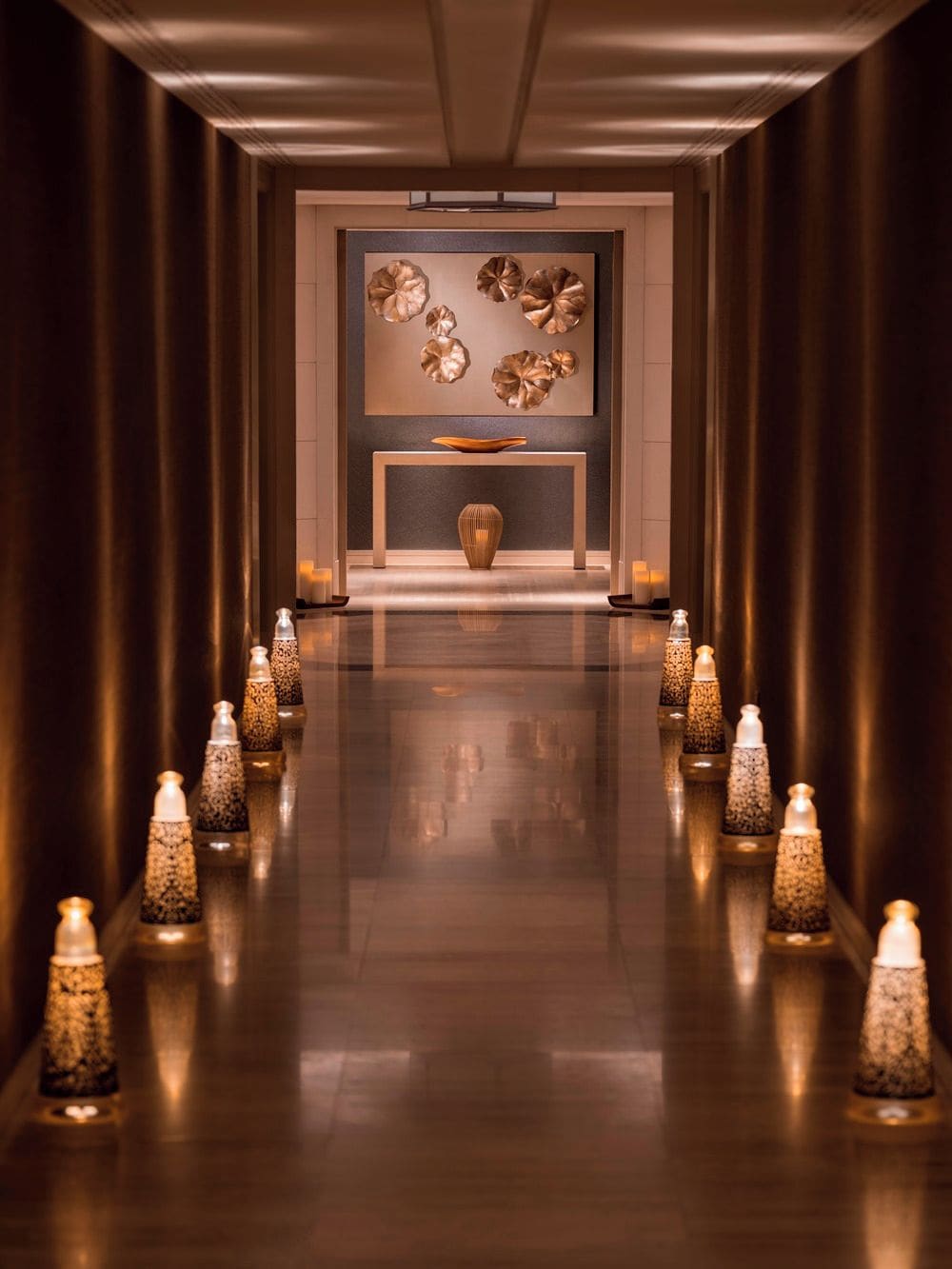 A corridor with a reflective floor leading to a reception area with a modern design, including a central table and decorative wall art.