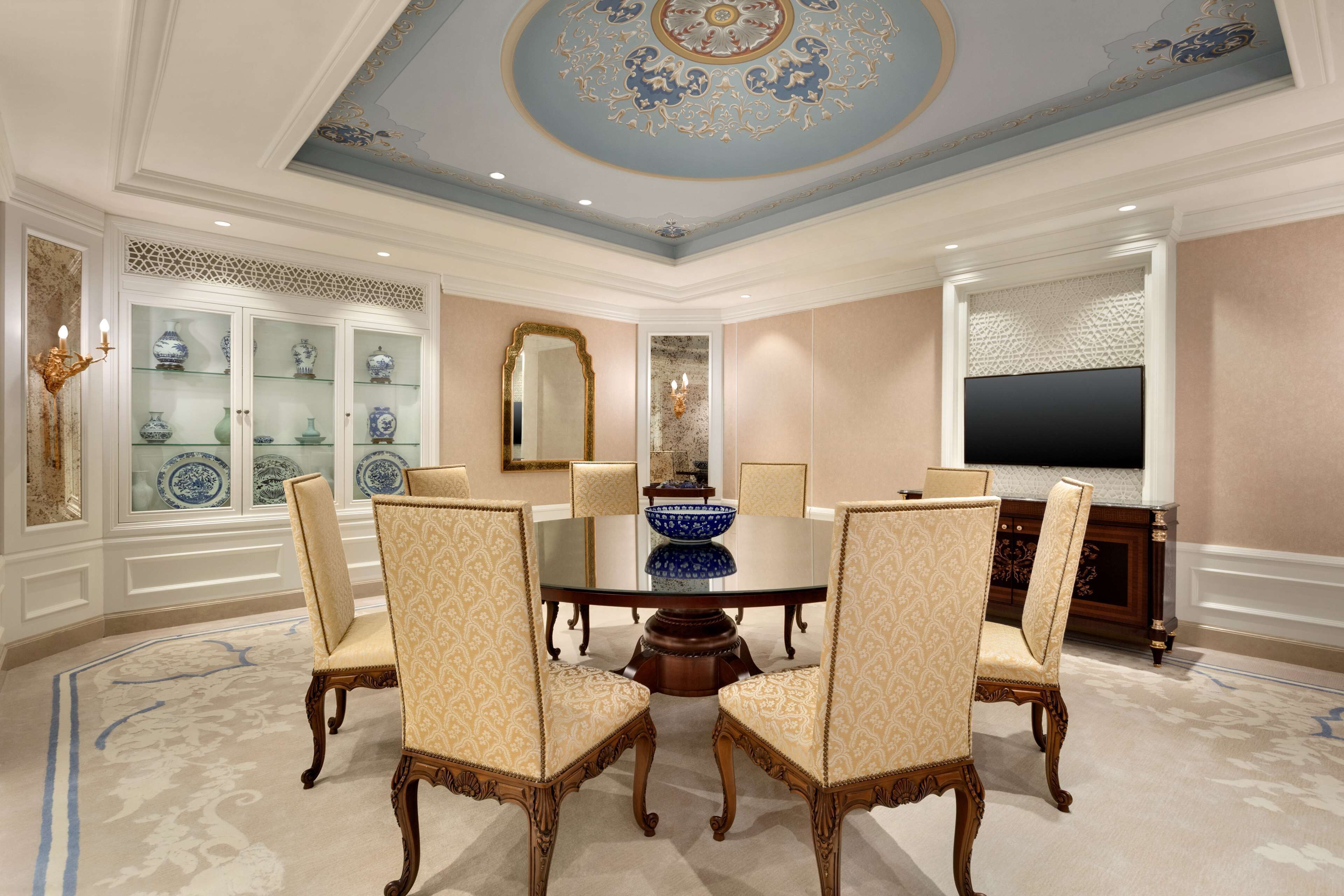 A formal dining room with a round table, six chairs, a decorative ceiling, and a television on the wall.