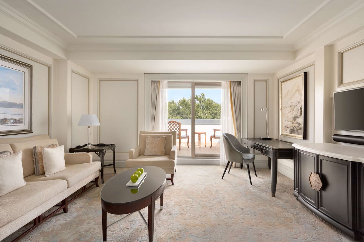 A hotel room with a sofa, a dining table, a TV, and a balcony with outdoor furniture.