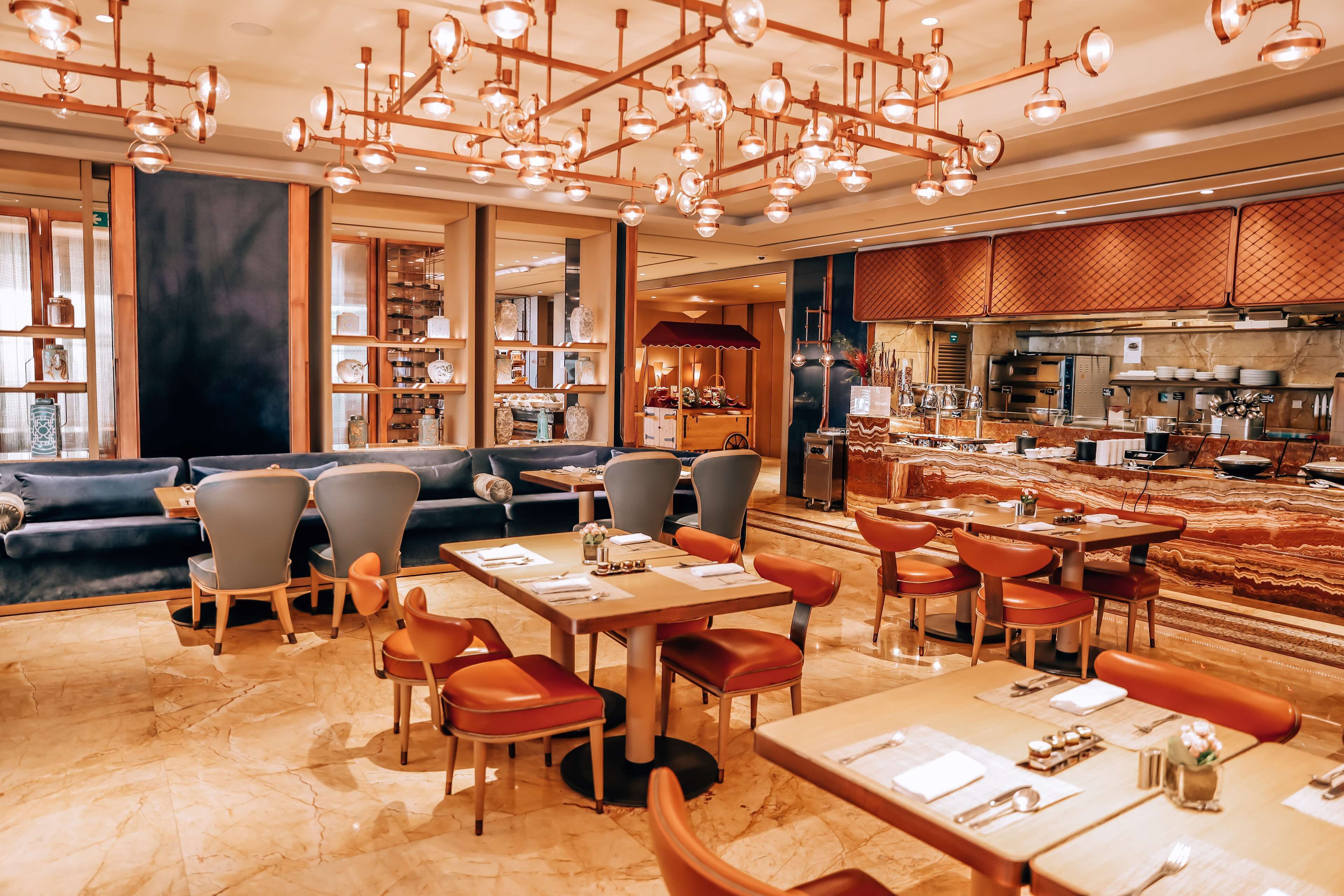 A restaurant with a modern interior, featuring a mix of seating options including booths and tables, a kitchen area visible in the background, and a chandelier with multiple light fixtures.