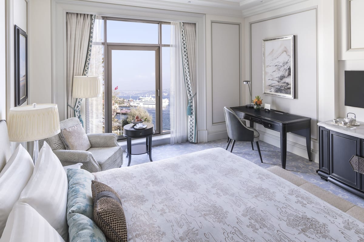 A hotel room with a large bed, a desk, a chair, a lamp, and a view of the city through large windows.