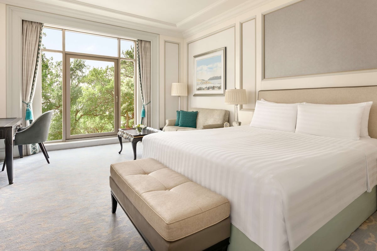 A hotel room with a large bed, a desk, a chair, and a view of trees through the window.