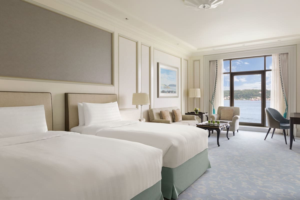 A hotel room with two large beds, a sitting area with a table and chairs, and a view of the sea through large windows.