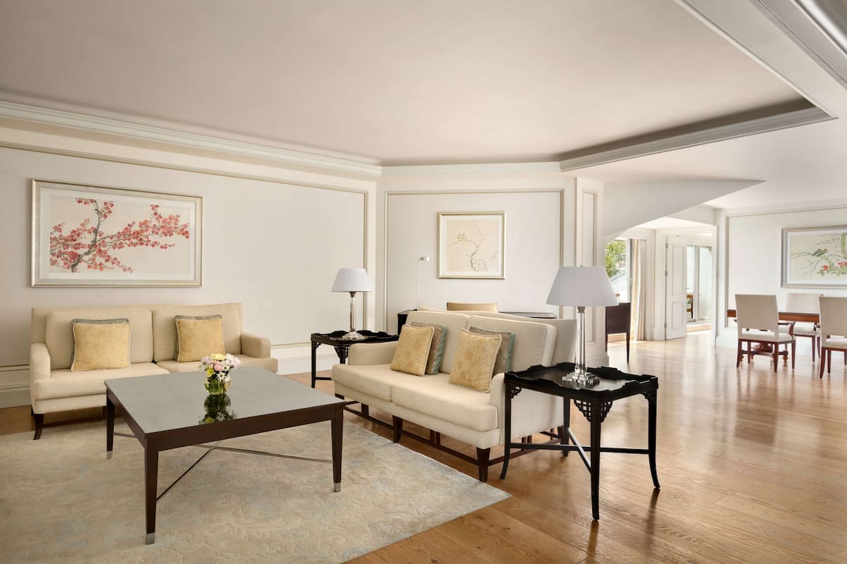 A spacious living room with a beige sofa, a coffee table, and a black side table. There are framed artworks on the walls and a wooden floor.