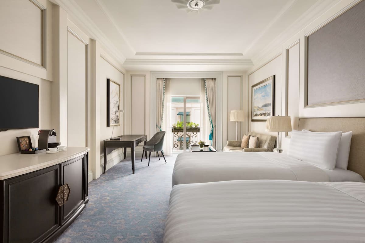 A hotel room with two beds, a desk, a chair, a TV, and a balcony with a view.