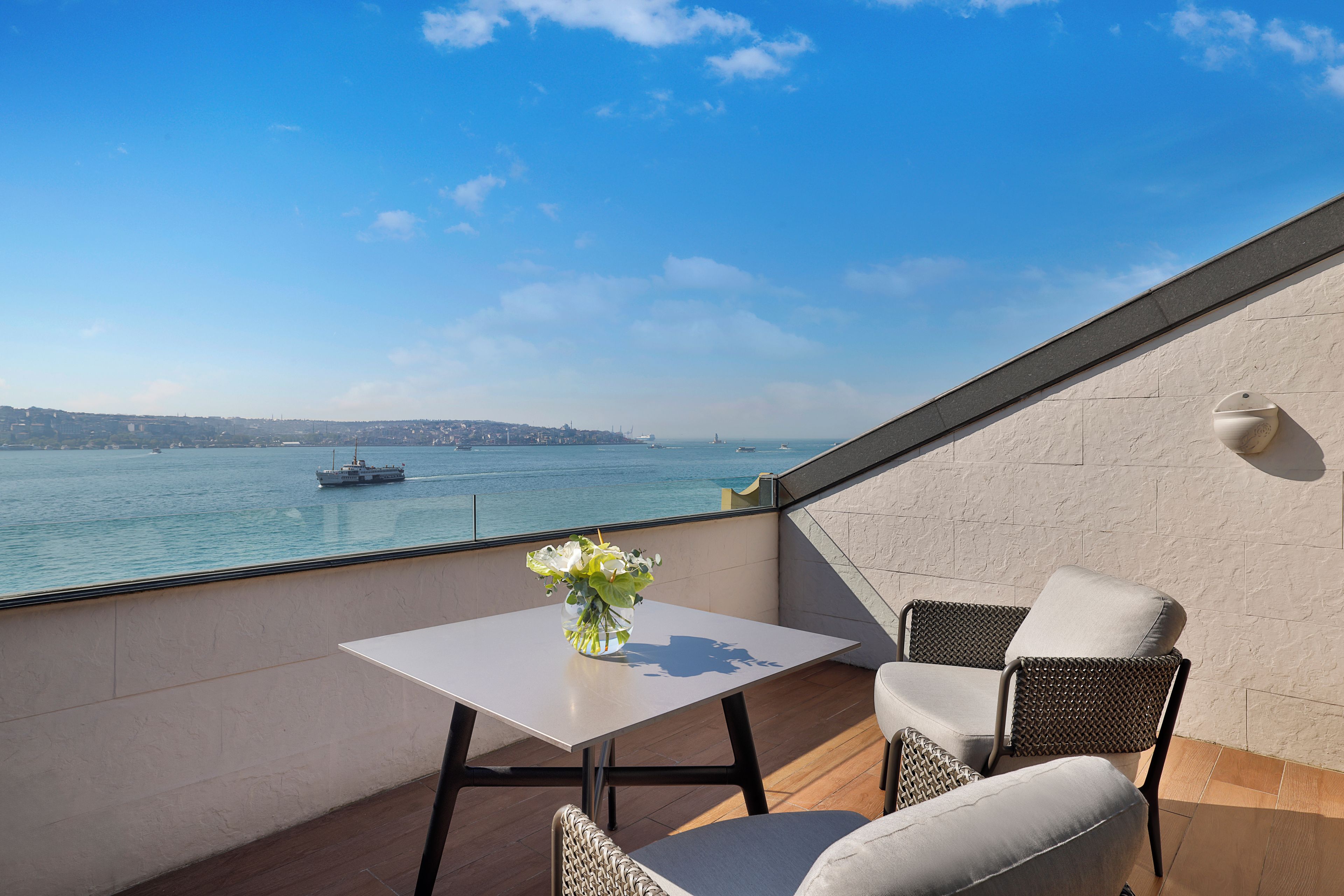 A balcony with a table and two chairs, overlooking a body of water with a ship in the distance.