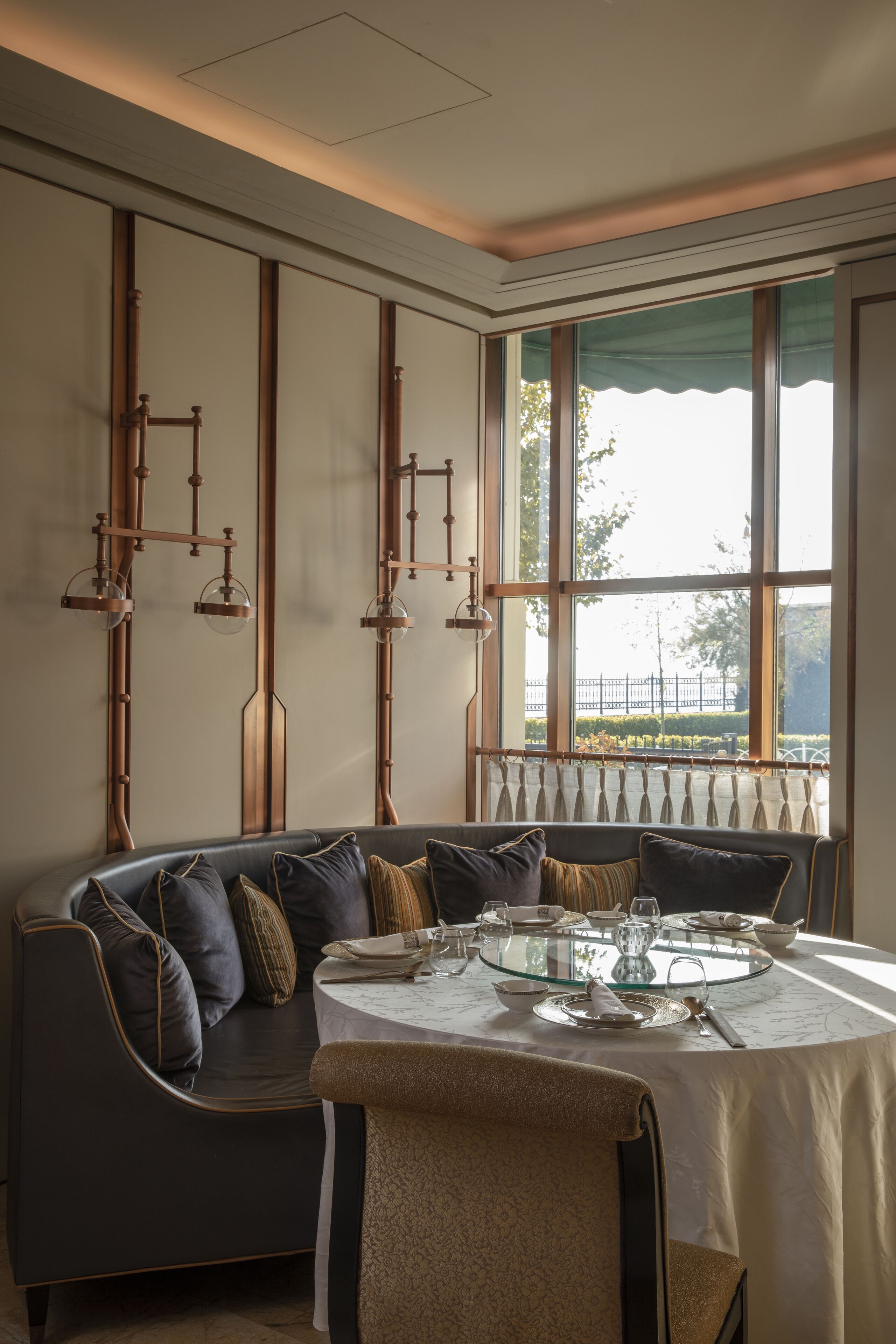 Un área de comedor con una mesa redonda dispuesta para cuatro personas, un sofá de cuero oscuro y una ventana con vistas a una valla y árboles.