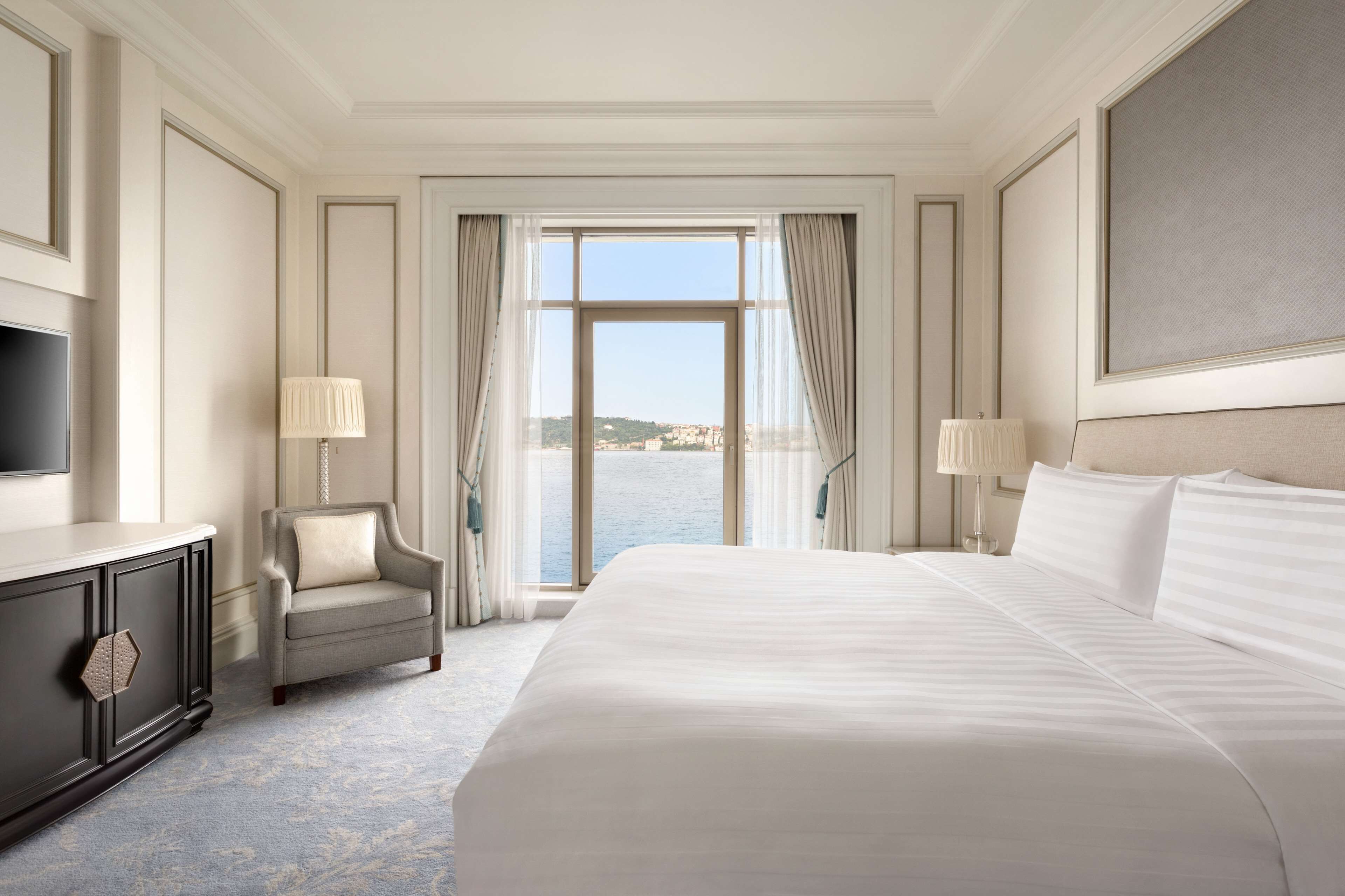 A hotel room with a large bed, a TV, a chair, and a view of the sea.