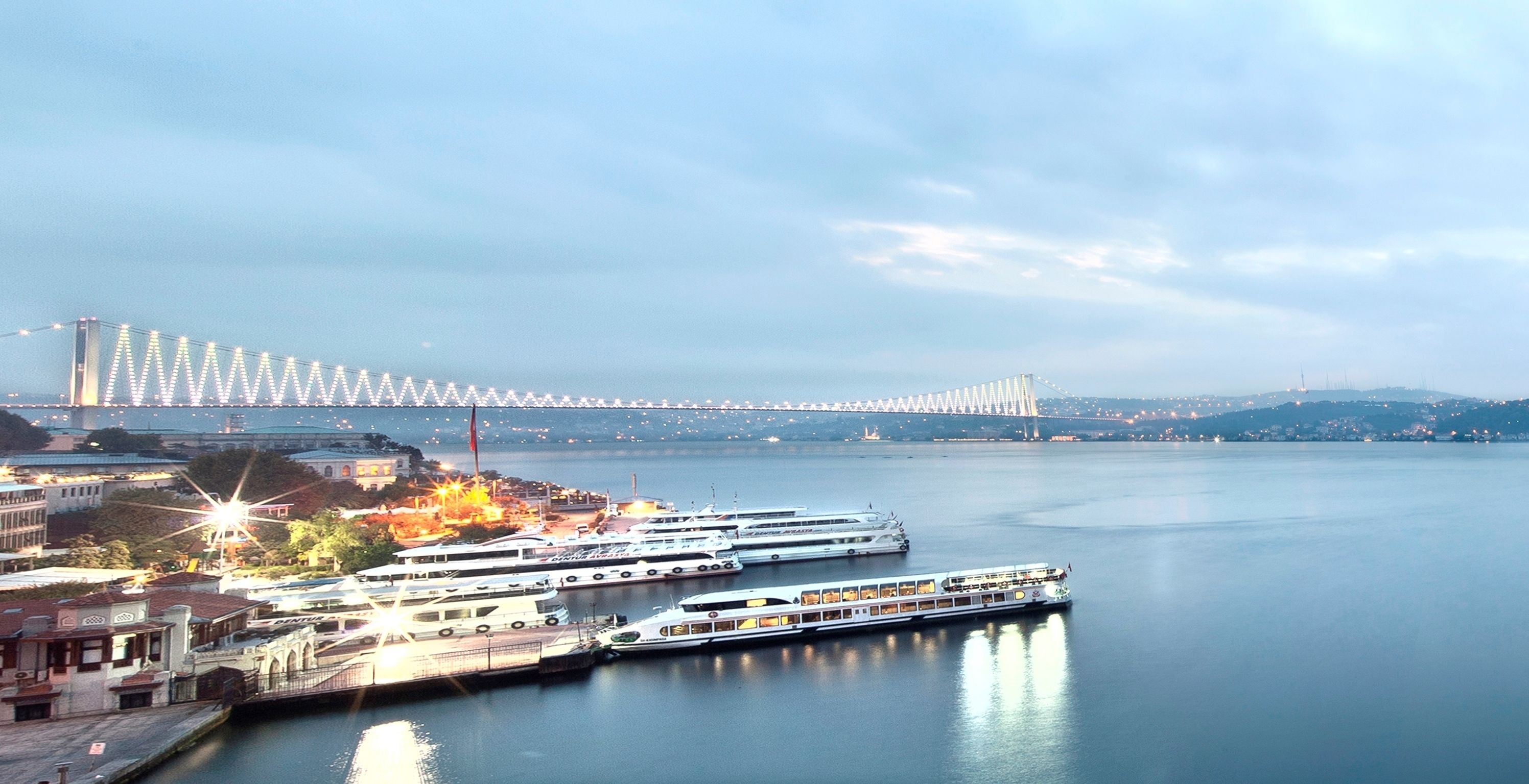 Un puerto deportivo frente al mar con varias embarcaciones amarradas, un puente grande al fondo y un paisaje urbano al otro lado del agua.