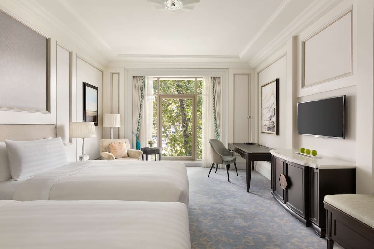 A hotel room with two beds, a desk, a chair, a TV, and a large window with curtains.