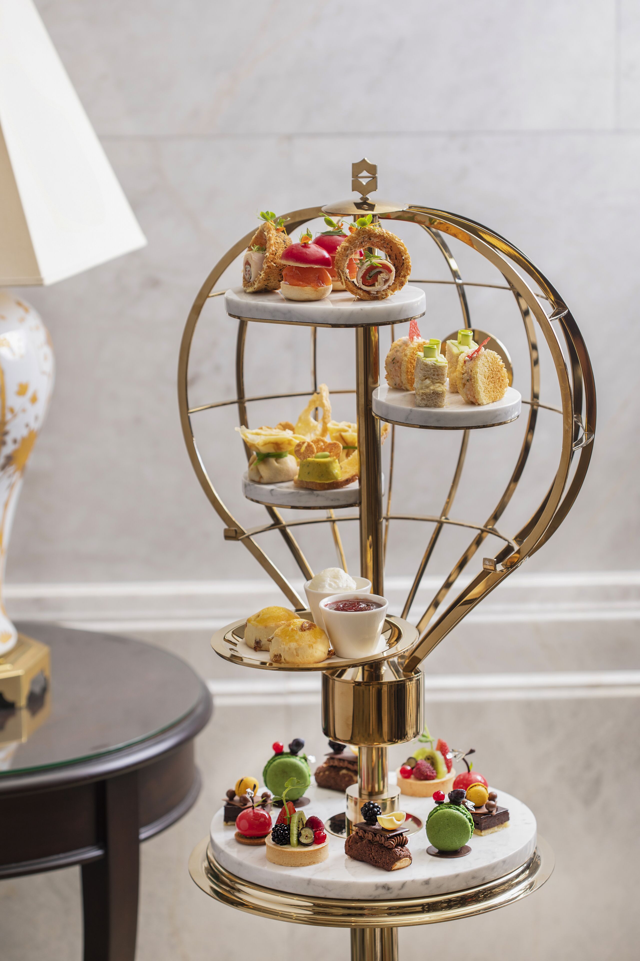 A multi-tiered dessert stand with various pastries and small cakes, set on a marble table against a neutral background.