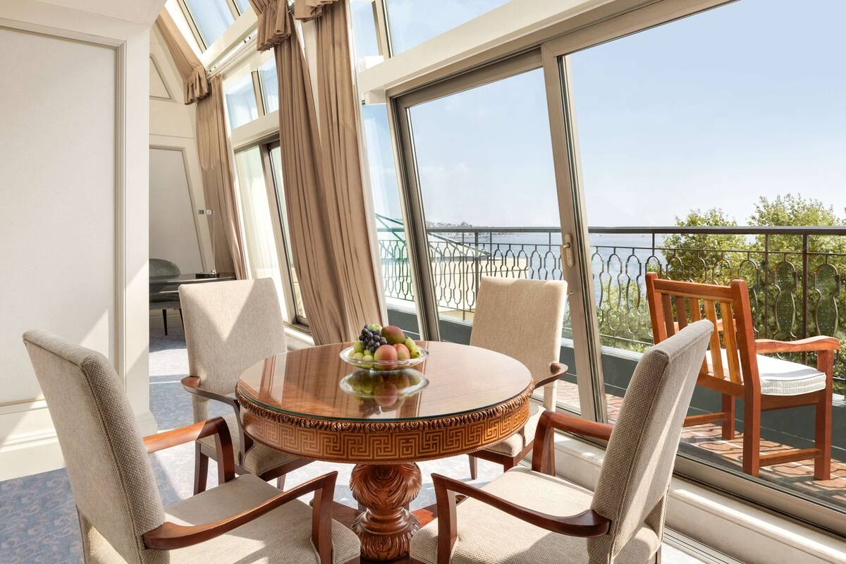 A round table with a fruit bowl, surrounded by four chairs, in a room with large windows overlooking a balcony with a railing and greenery.