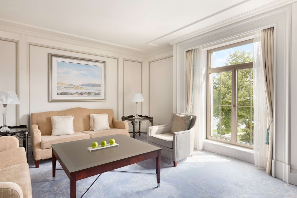 A spacious living room with a beige sofa, a matching armchair, a wooden coffee table with apples, and a large window with curtains.