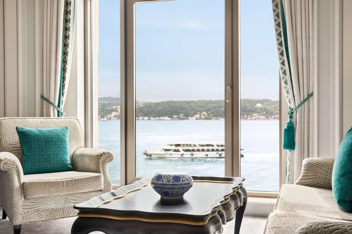 A room with a view of a boat on the water, a chair with a teal pillow, and a table with a blue and white bowl.