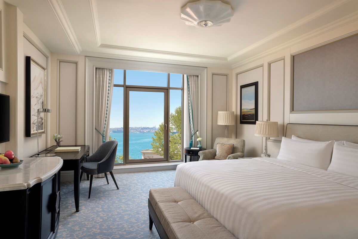A hotel room with a large bed, a desk, a chair, a view of the sea, and a cityscape.