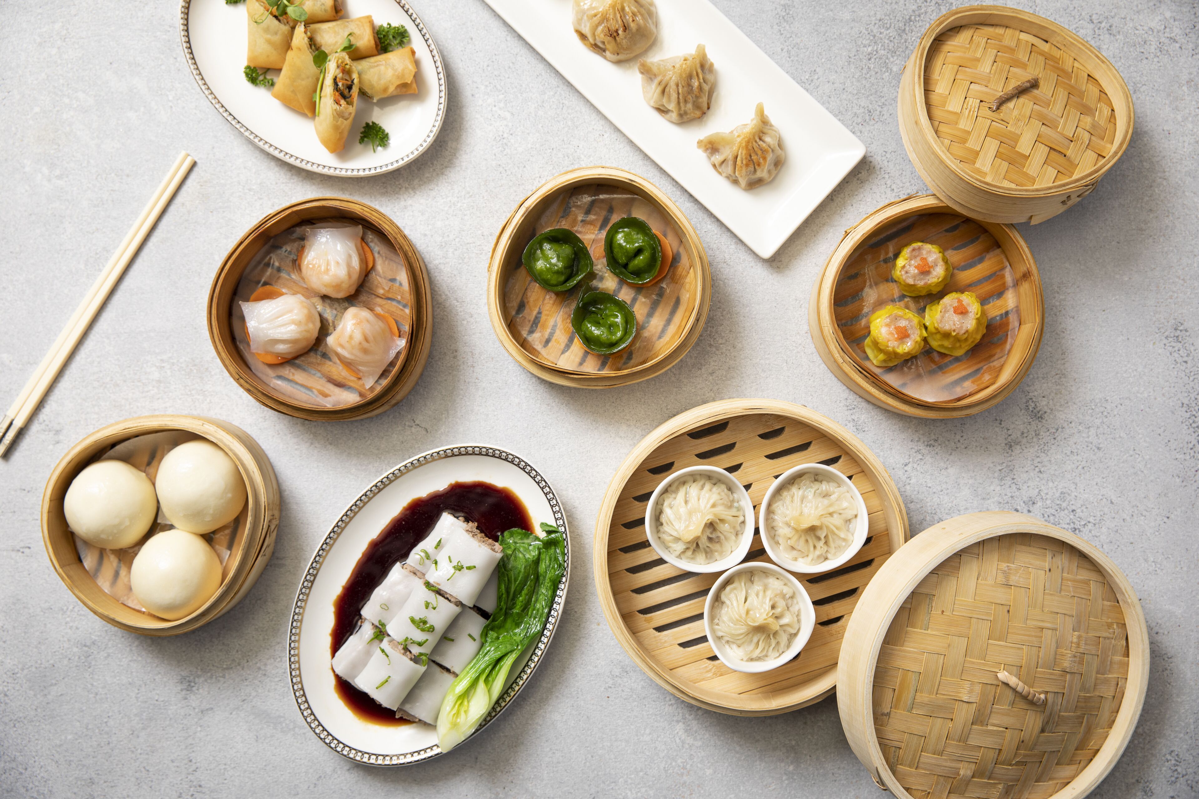 A variety of Asian dumplings and buns in bamboo steamers, some with dipping sauces, and a pair of chopsticks on a table.
