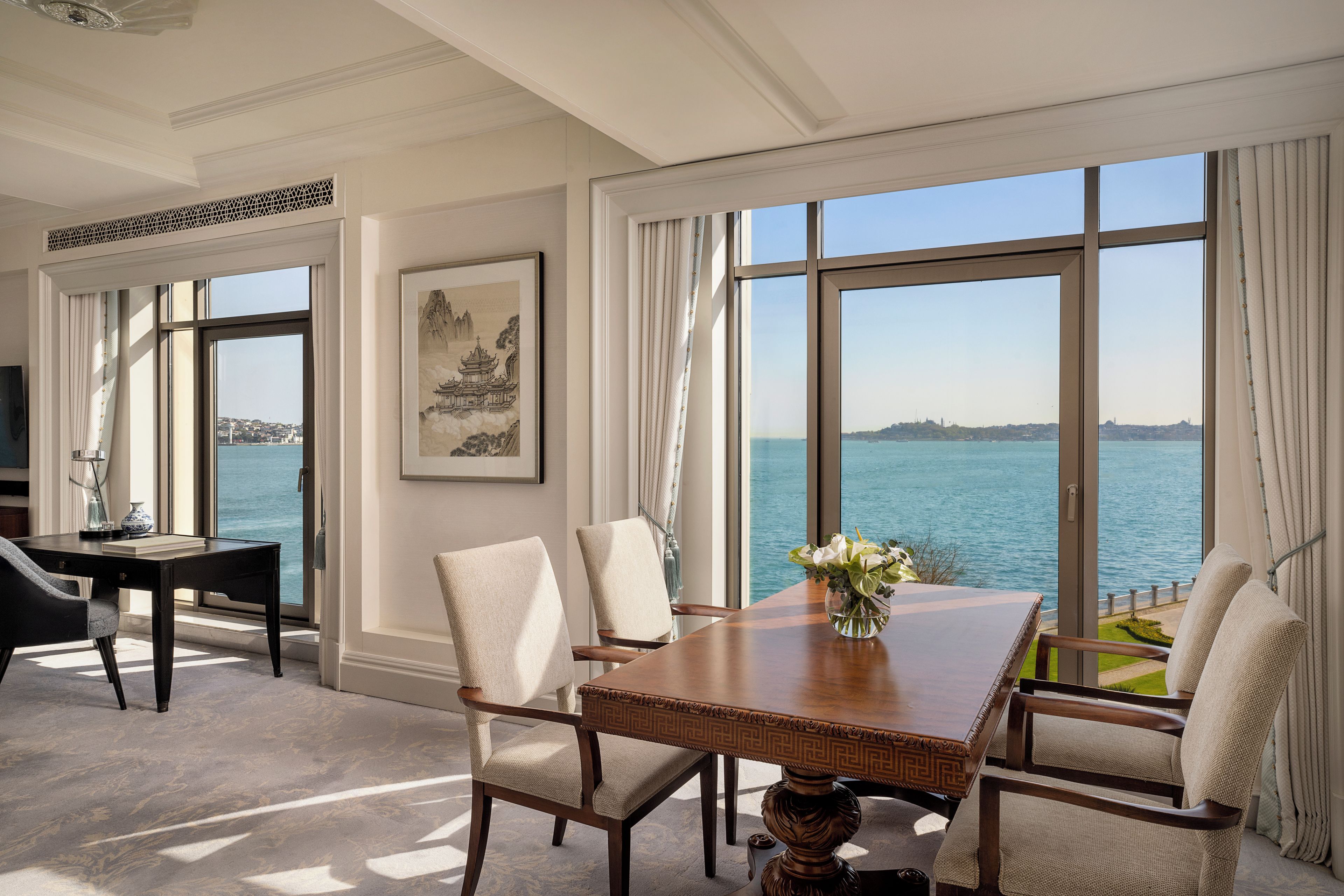 Un comedor con mesa y sillas, ventanas amplias con vista al mar y una obra de arte enmarcada en la pared.