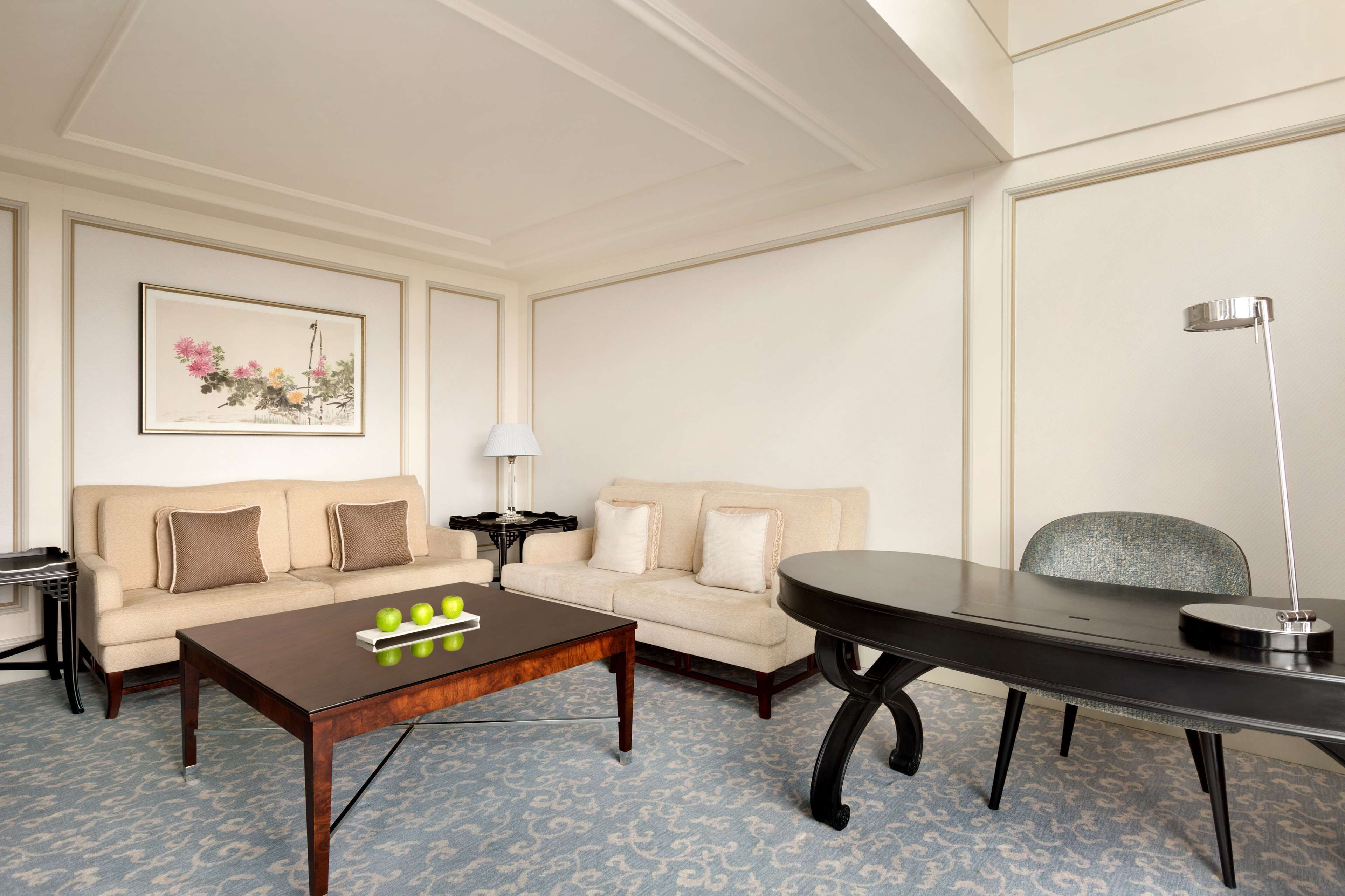 A room with a beige sofa, a wooden coffee table with apples, a black desk, and a lamp.
